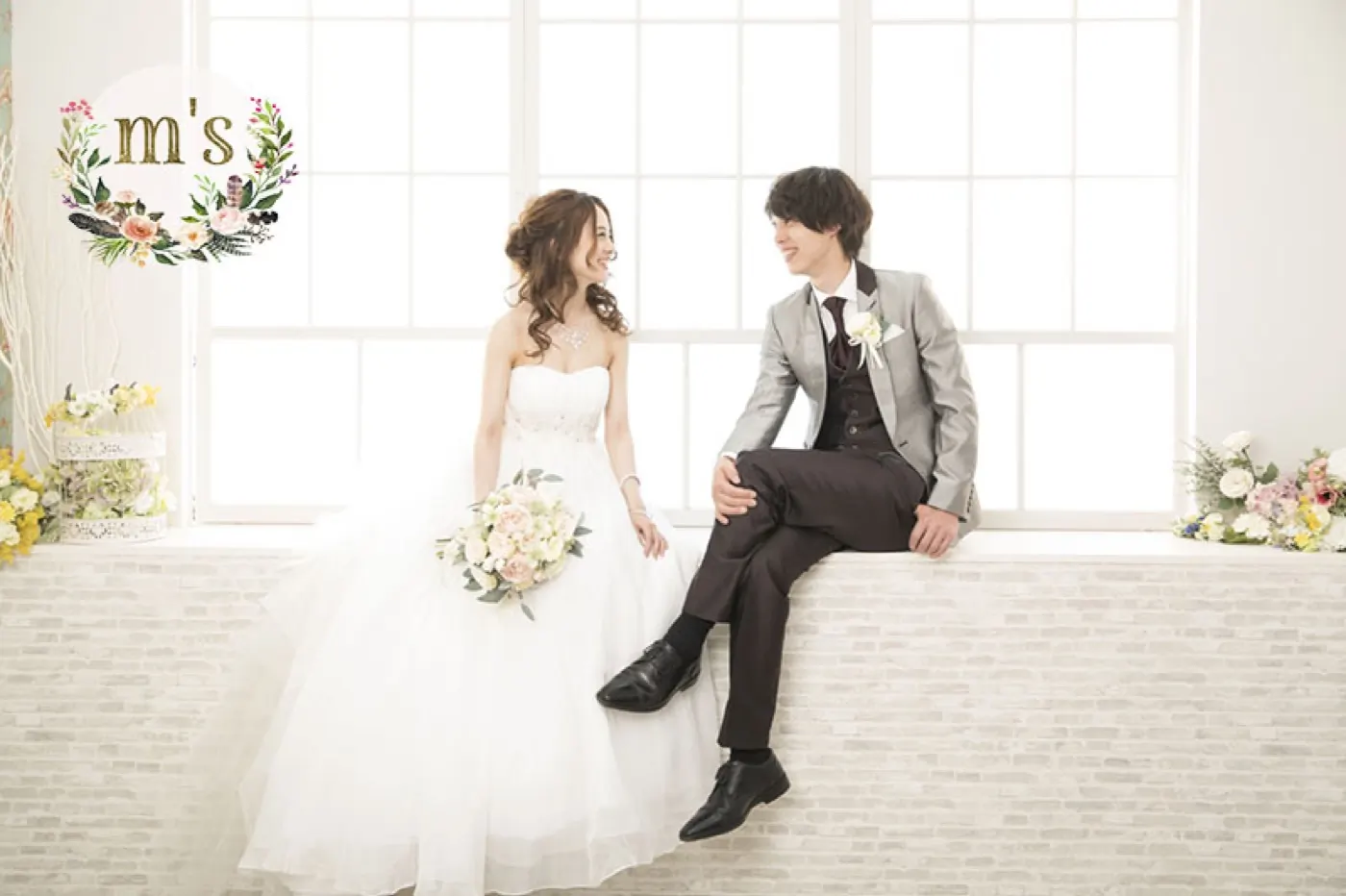 窓辺の明るいスタジオで並んで座る、ドレスとタキシードの新郎新婦