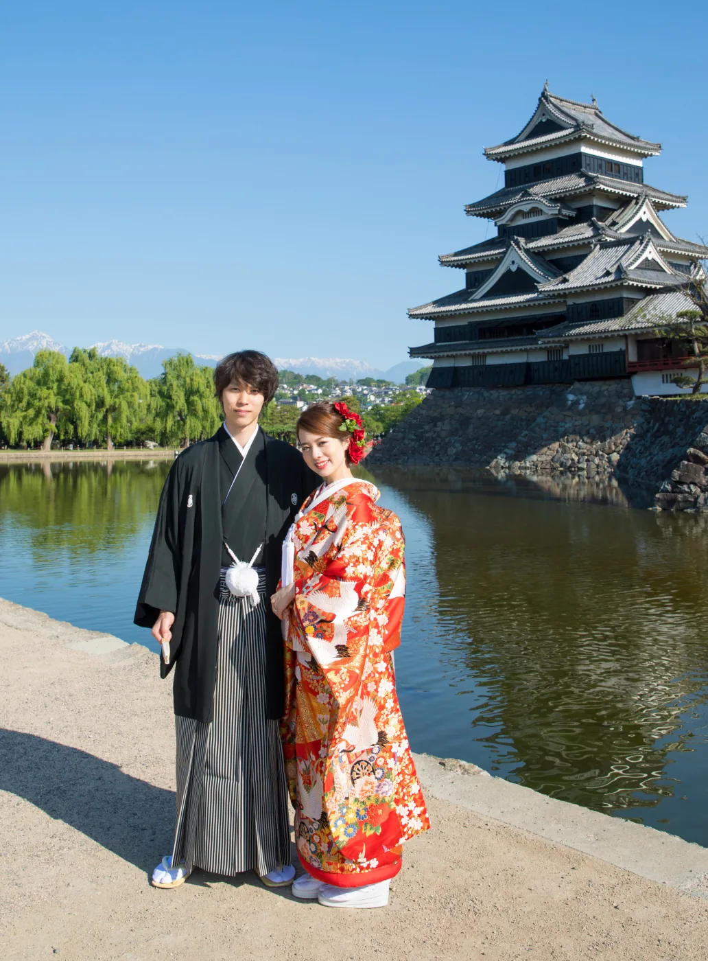 青空の下、松本城の天守と堀を背に並ぶ赤い色打掛の花嫁と黒紋付袴の花婿。左奥に雪をかぶった北アルプスが見える