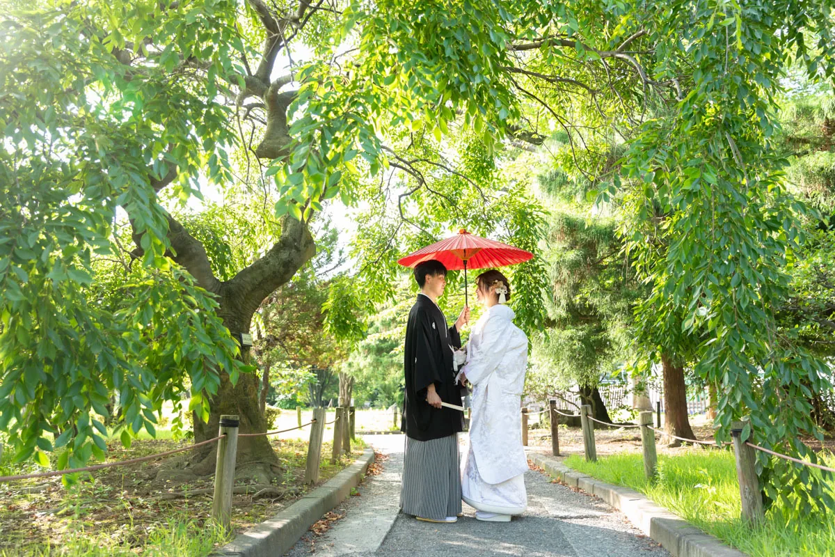緑のトンネルの下で赤い和傘をさす、白無垢の花嫁と黒紋付袴の花婿