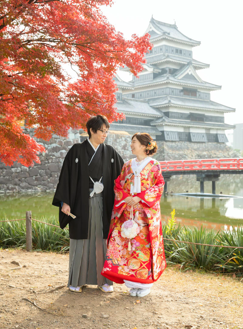真っ赤なもみじ越しに松本城の天守と赤い埋橋を望んで並ぶ、赤い色打掛の花嫁と黒紋付袴の花婿