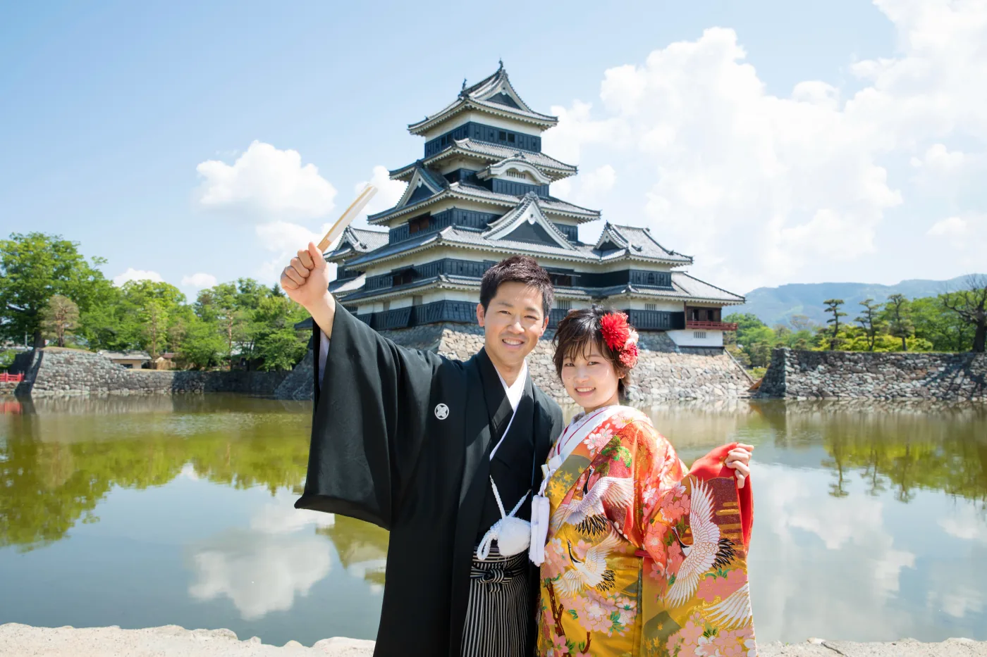 青空と松本城の天守、堀を背景に、扇子を高くかかげて笑う黒紋付袴の花婿と色打掛の花嫁