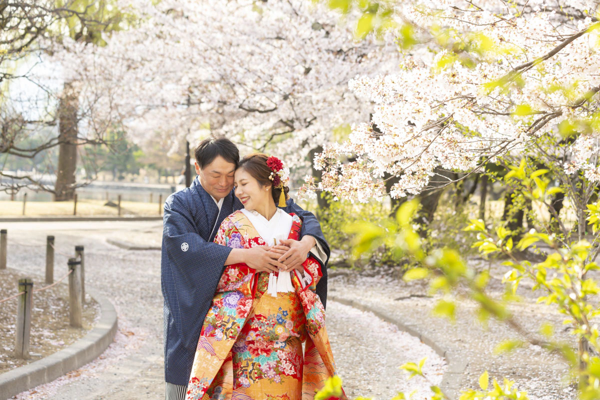 散りはじめの桜の下で寄り添う、色打掛の花嫁と紺の羽織袴の花婿