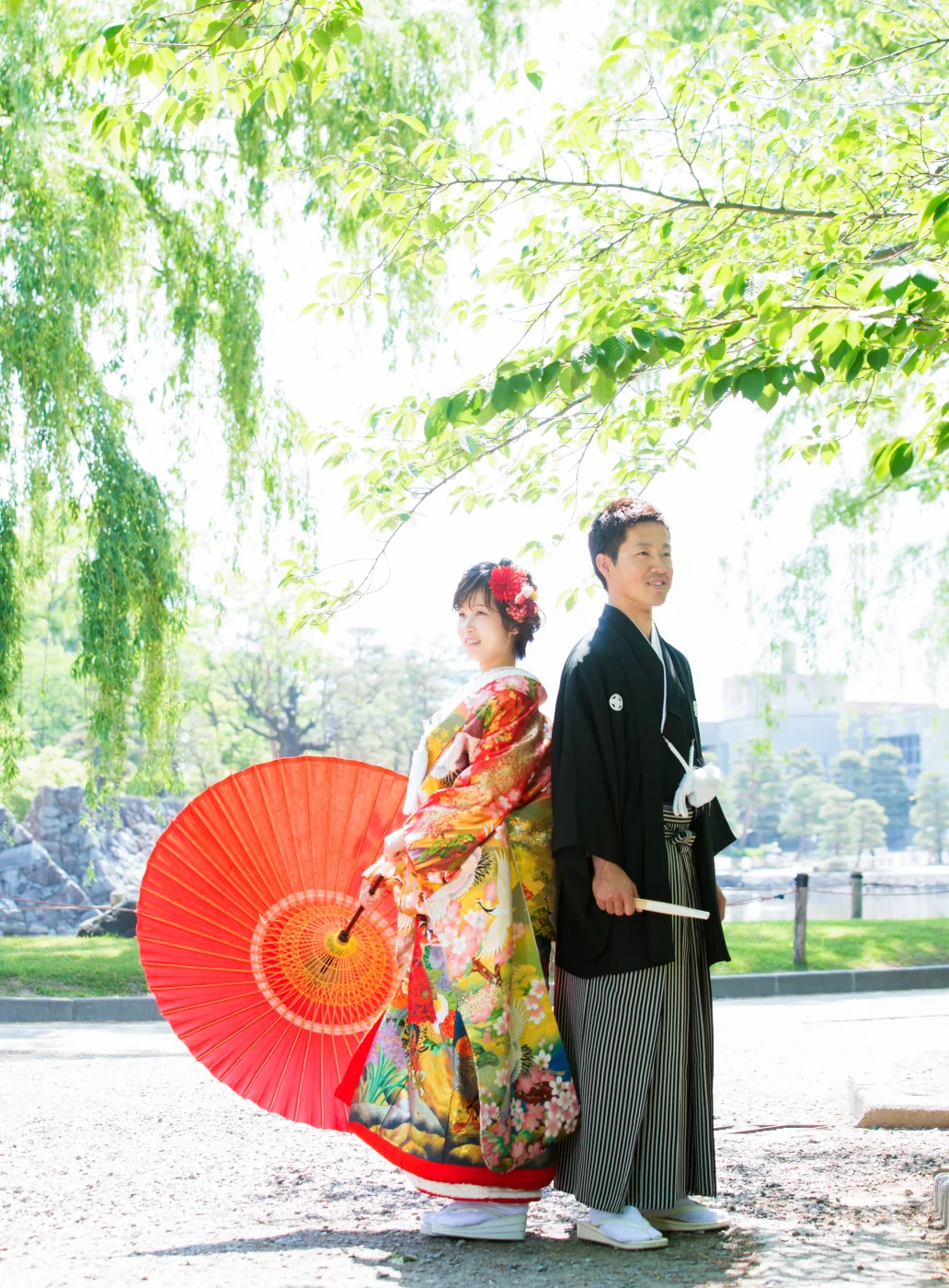 新緑の柳の下で背中合わせに立つ、赤い和傘を持った色打掛の花嫁と黒紋付袴の花婿