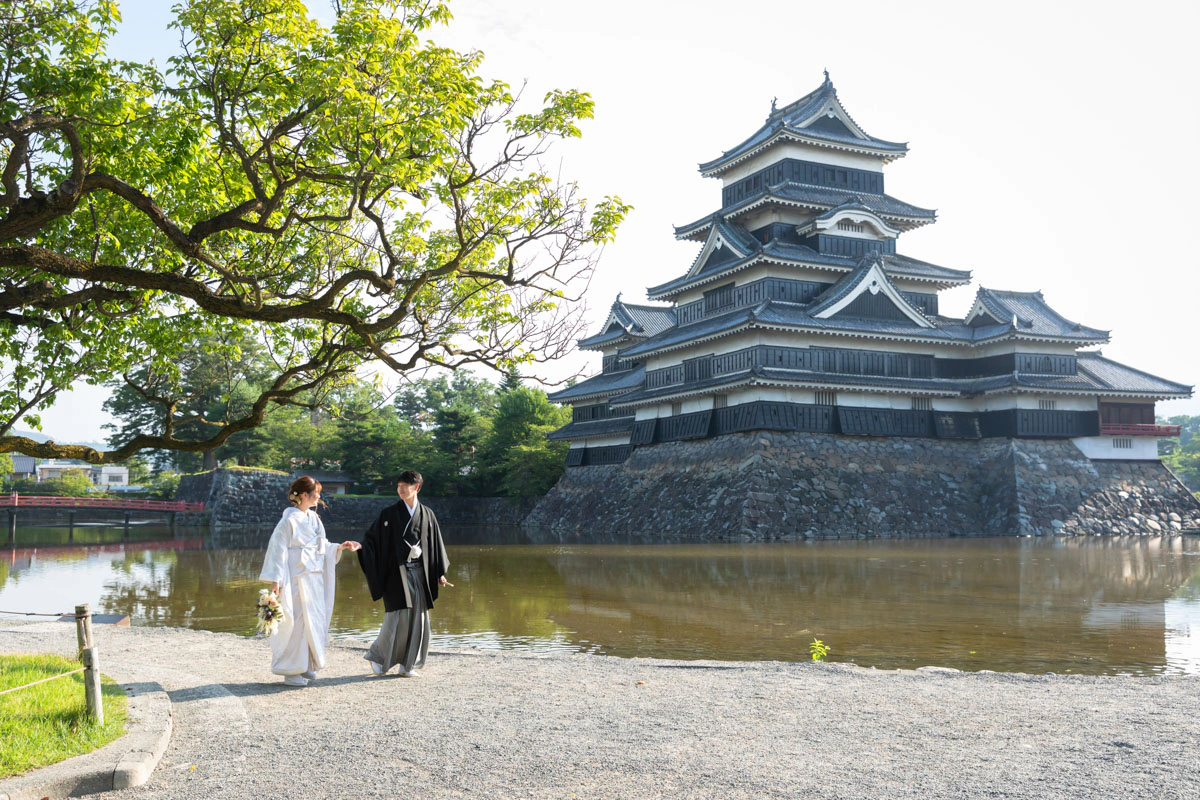 朝の松本城の天守と堀を背景に、堀端を手をつないで歩く白無垢の花嫁と黒紋付袴の花婿