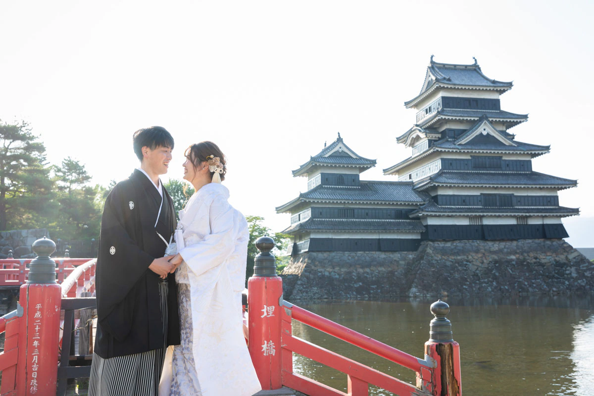 朝の松本城の天守を背に、赤い埋橋のたもとで向かい合う白無垢の花嫁と黒紋付袴の花婿