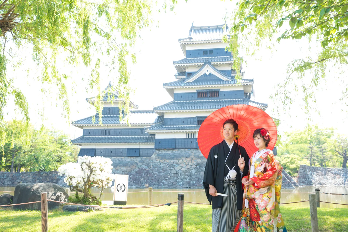柳の新緑ごしに松本城の天守を背景にして、赤い和傘をさして並ぶ色打掛の花嫁と黒紋付袴の花婿
