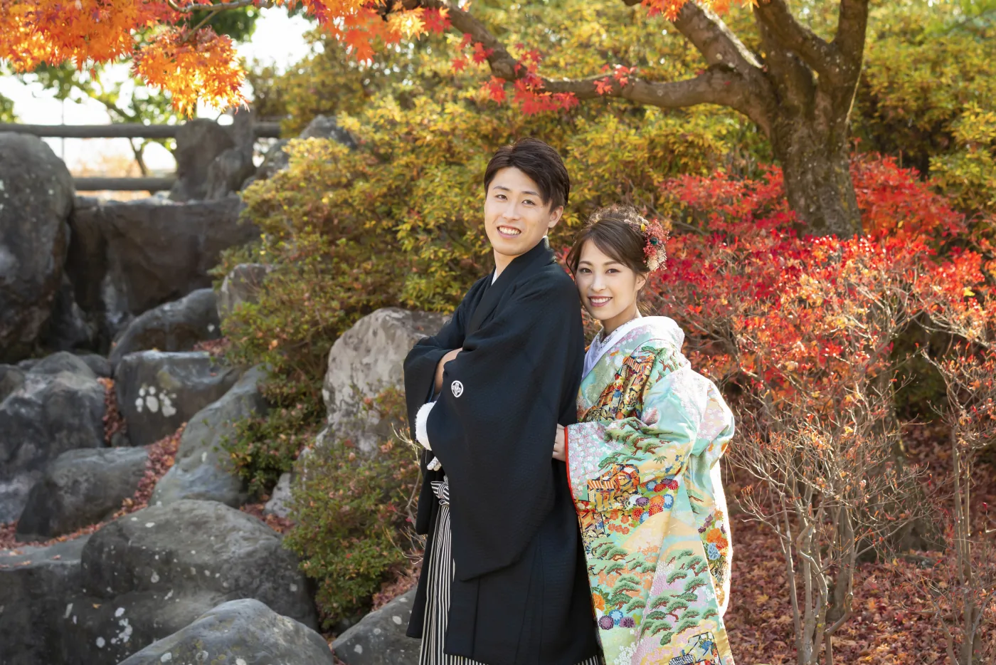 あがたの森の紅葉の下、岩を背に寄り添って立つ色打掛の花嫁と黒紋付袴の花婿
