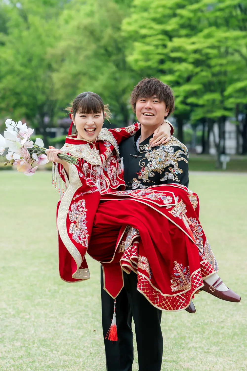 新緑の公園の芝生で、花婿にお姫様抱っこをされて笑う、赤い中華風の衣装の花嫁