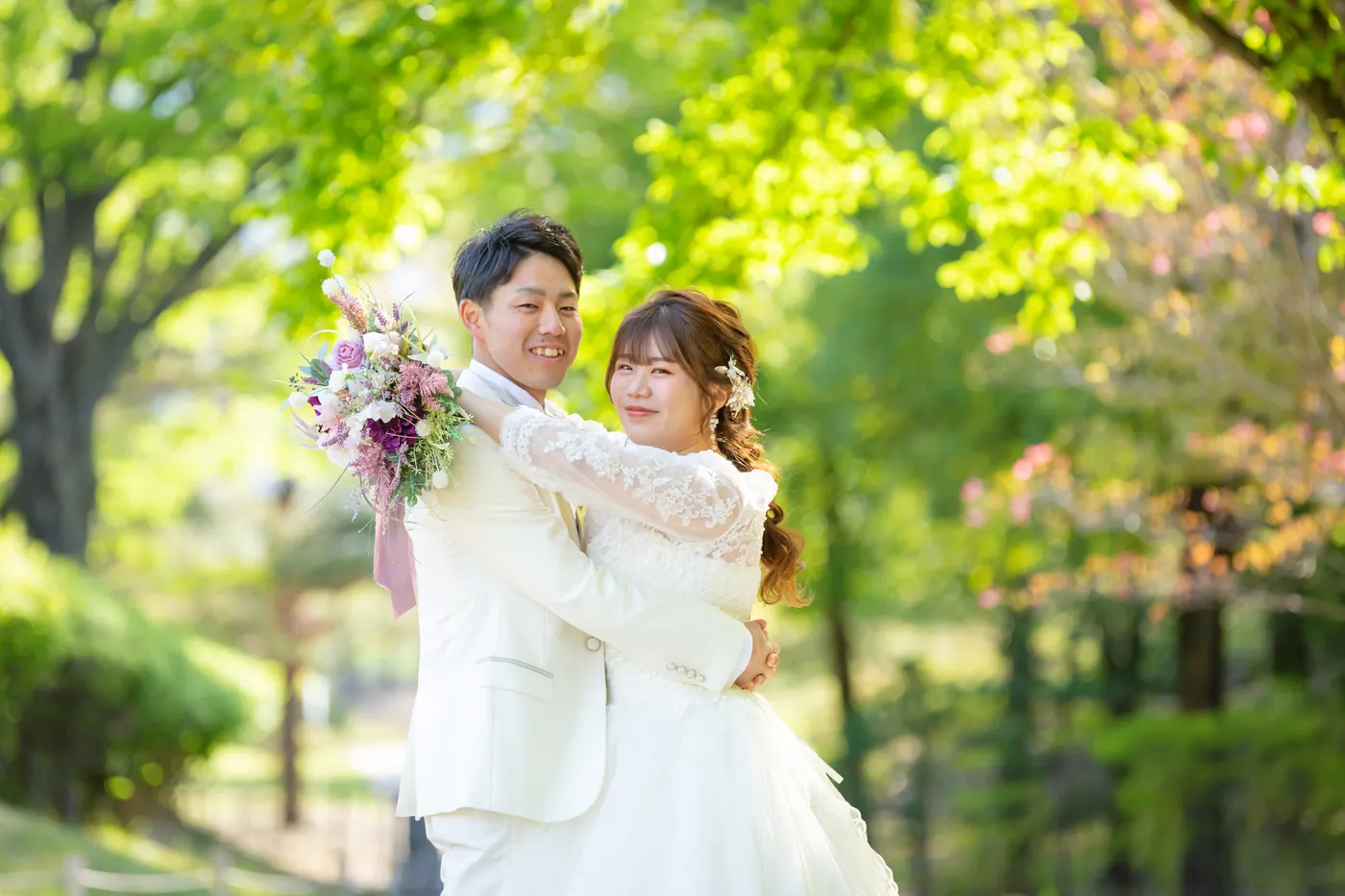 新緑の中でブーケを持ち寄り添う、ウェディングドレスの花嫁と白いタキシードの花婿
