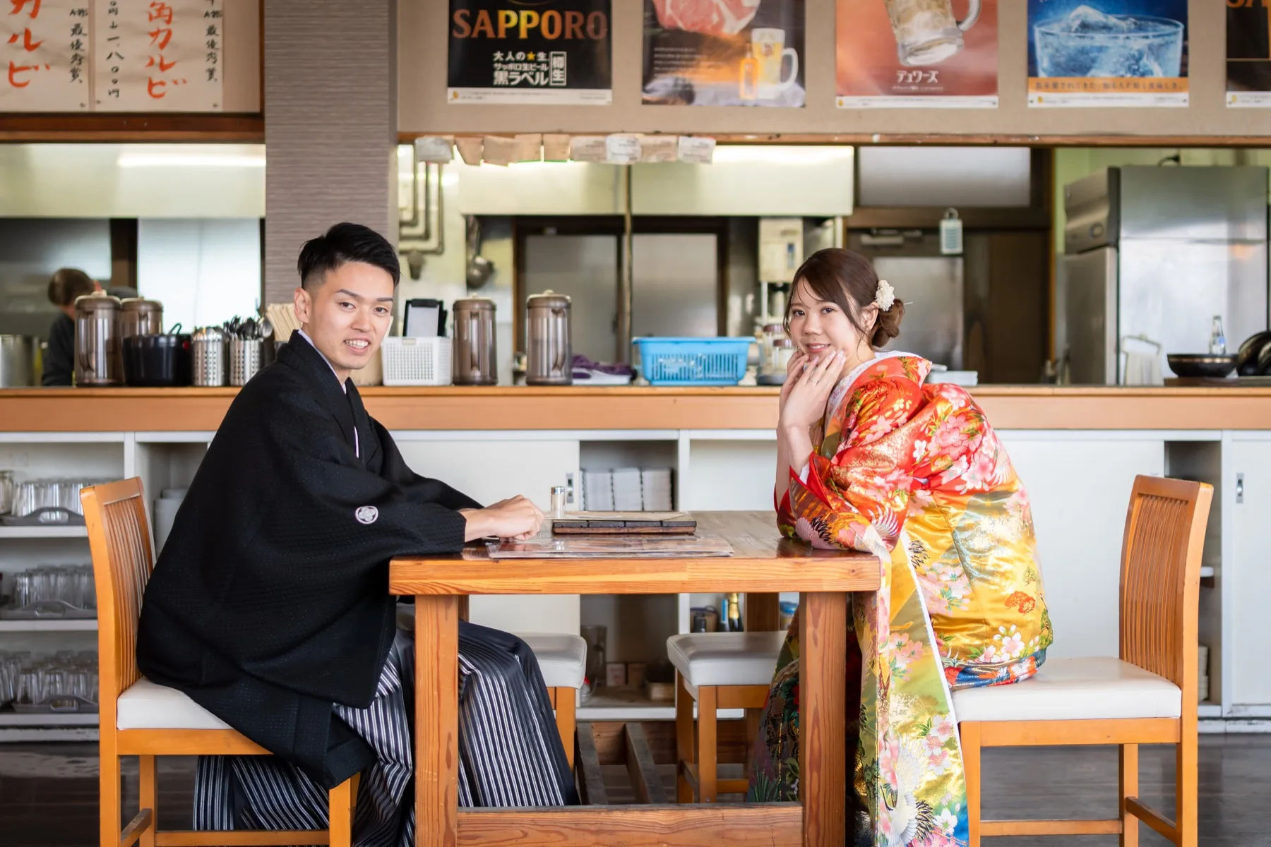 お二人の思い出の焼肉店のテーブル席で、向かい合って座る色打掛の花嫁と黒紋付袴の花婿