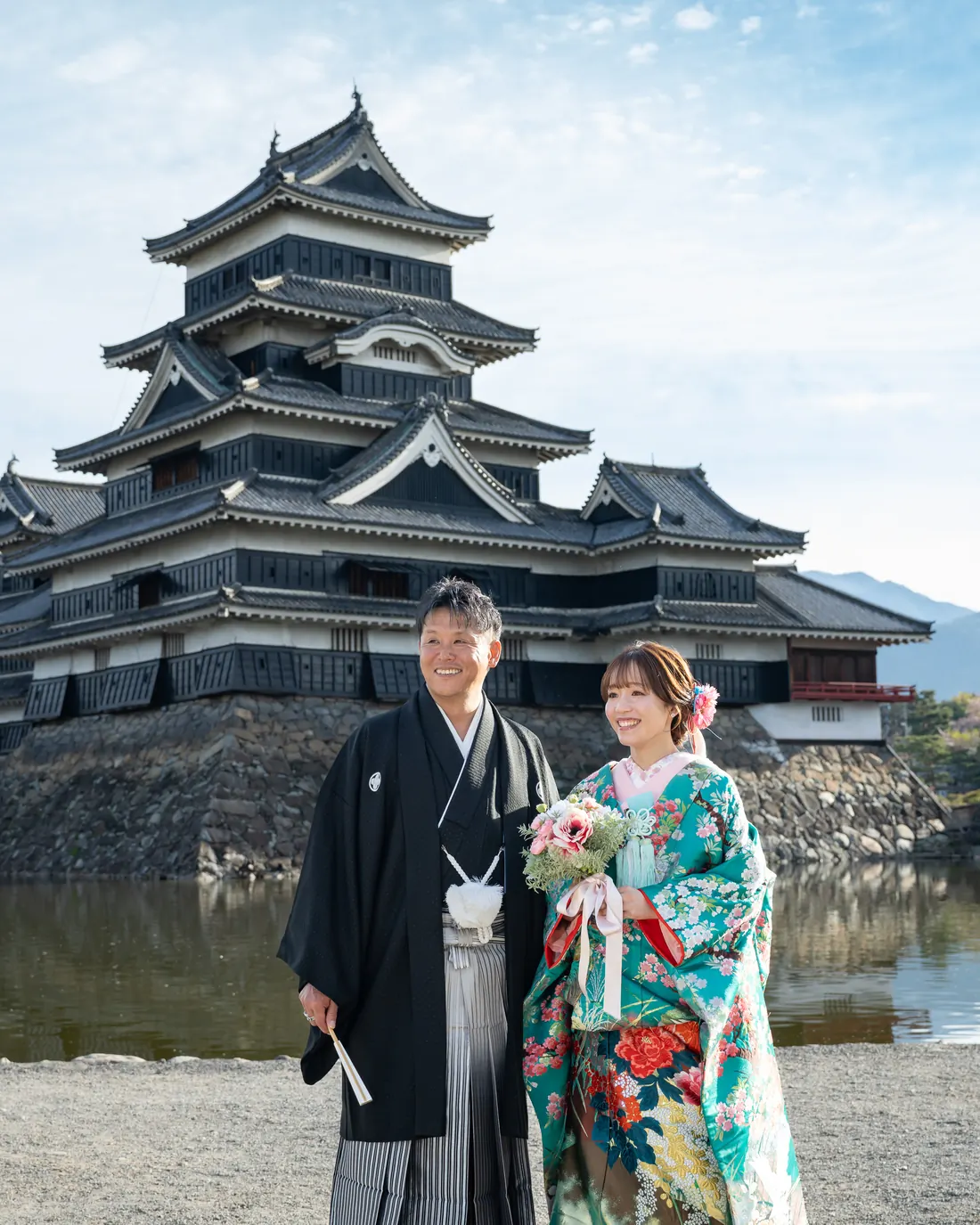 松本城と堀を背景に寄り添う、緑の色打掛の花嫁と黒紋付袴の花婿