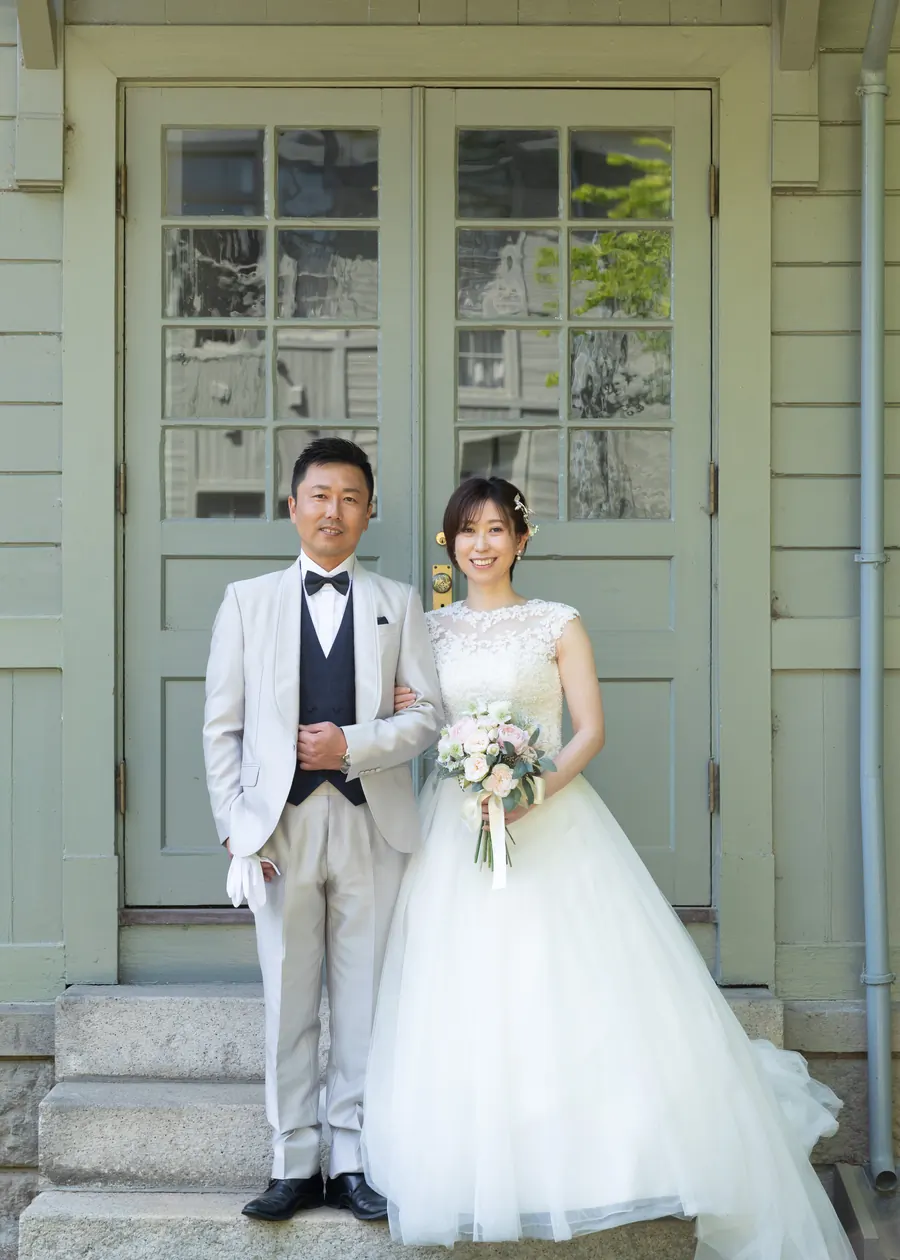 洋館の扉の前に立つ、ウェディングドレスの花嫁と白いタキシードの花婿