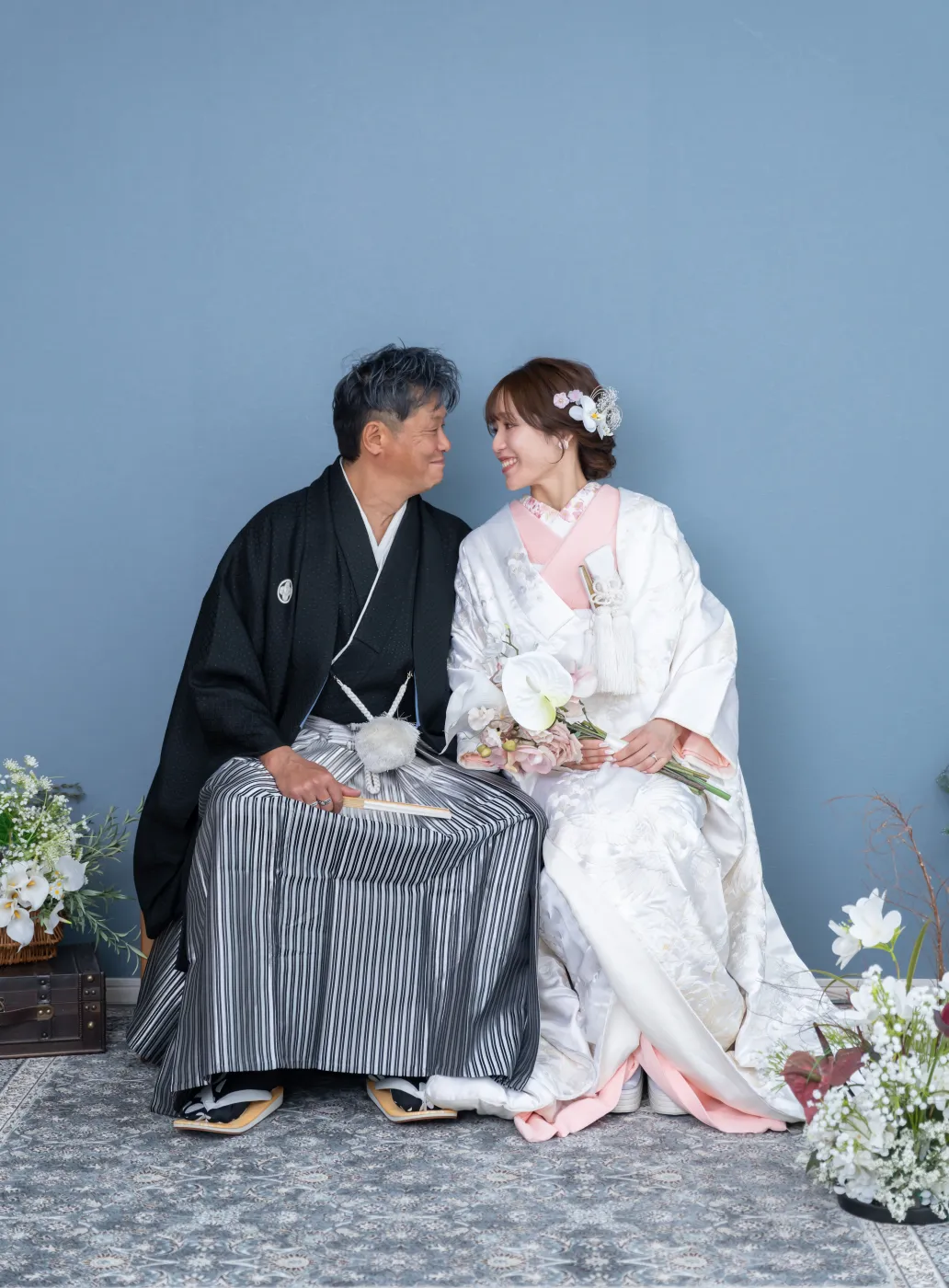 青い壁の前に並んで座り、見つめ合う白無垢の花嫁と黒紋付袴の花婿