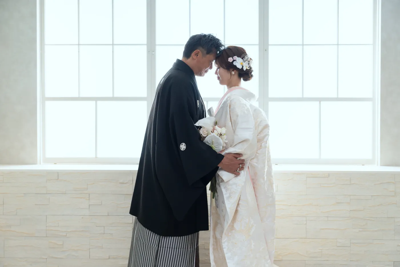 大きな窓の前で、額を寄せ合う白無垢の花嫁と黒紋付袴の花婿