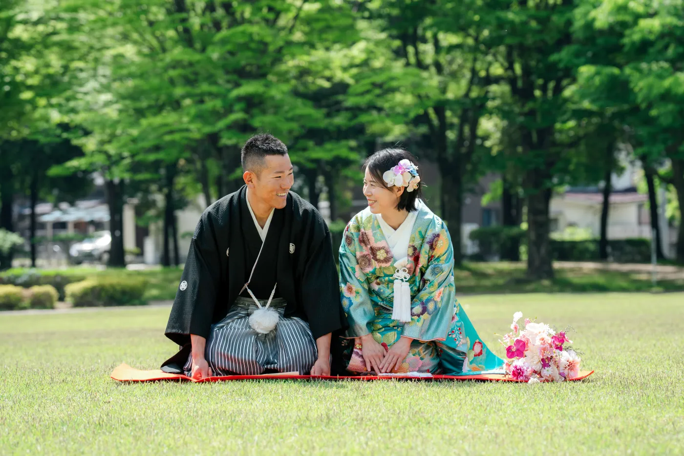 新緑の芝生に座り、顔を見合わせて笑う水色の色打掛の花嫁と黒紋付袴の花婿
