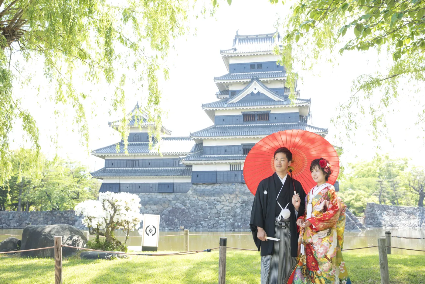 柳の新緑ごしに松本城の天守を背景にして、赤い和傘をさして並ぶ色打掛の花嫁と黒紋付袴の花婿