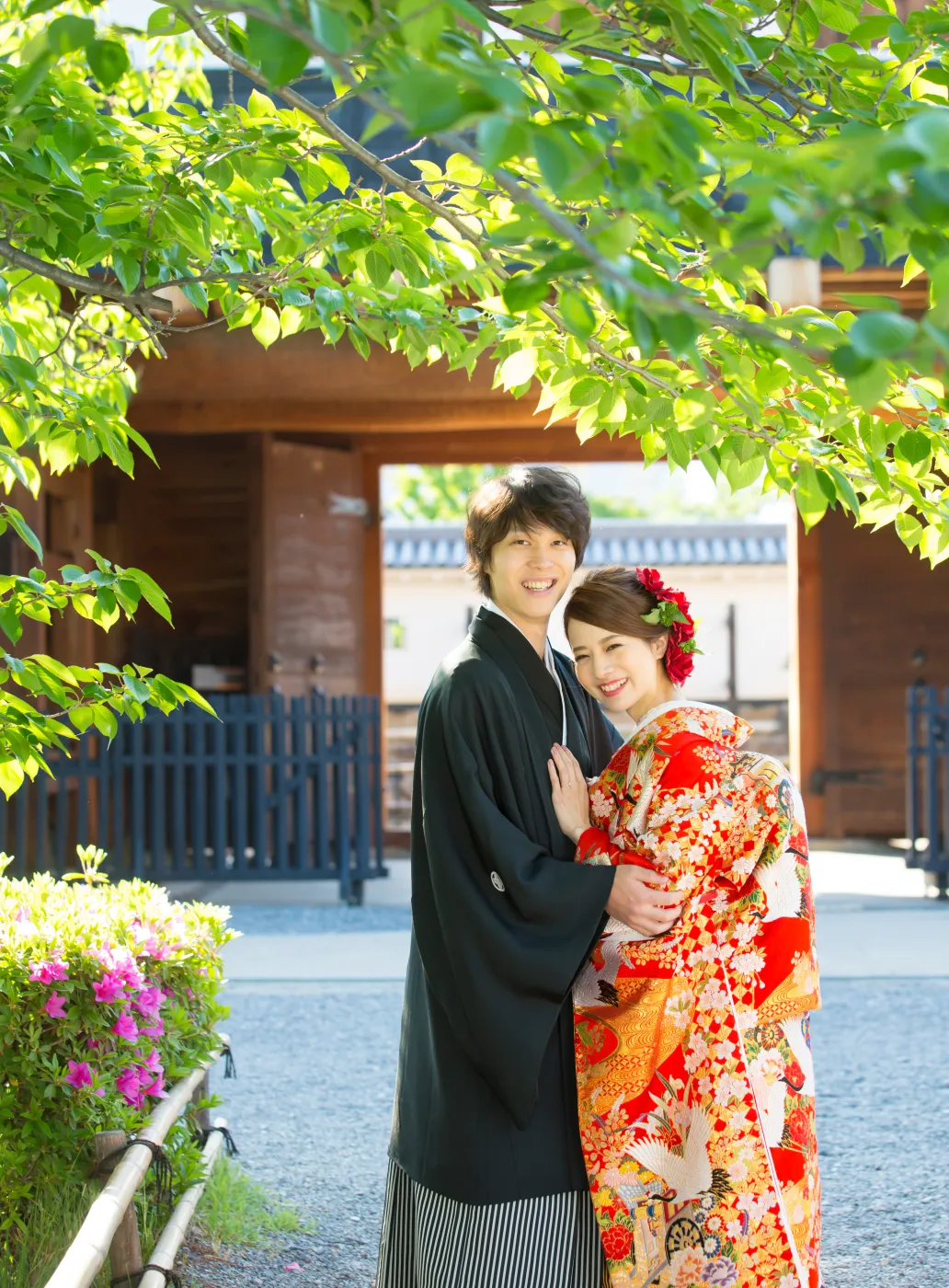 木の門の前で、笑顔で寄り添う赤い色打掛の花嫁と黒紋付袴の花婿
