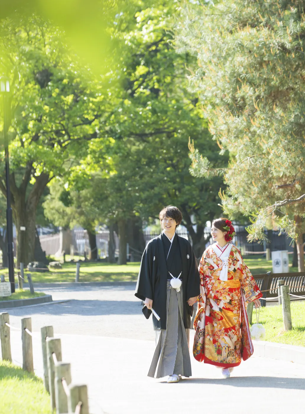 新緑の公園の道を、手をつないで歩く赤い色打掛の花嫁と黒紋付袴の花婿