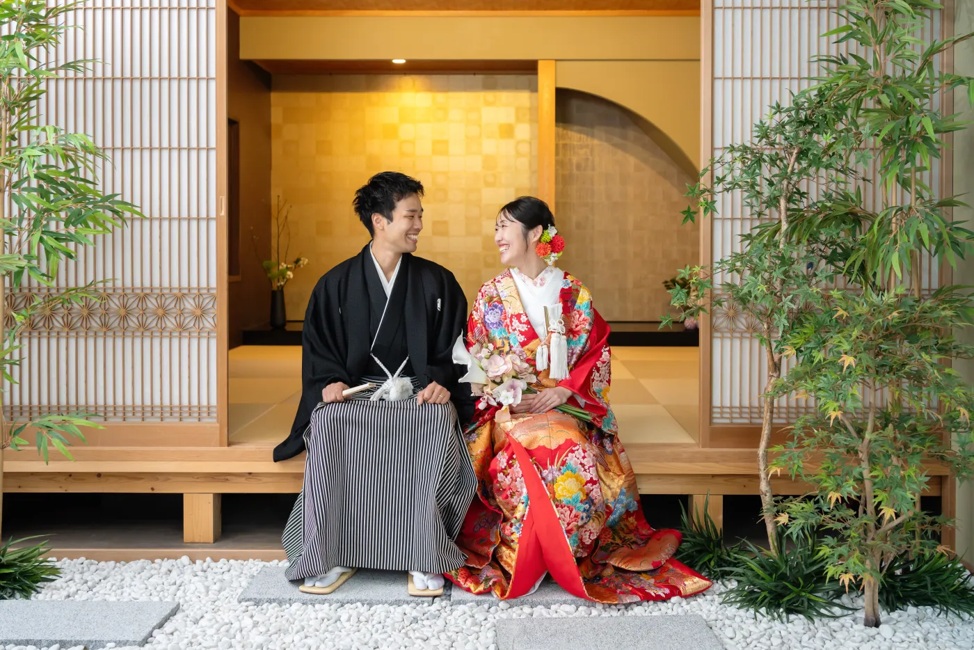 和室の縁側に並んで座り、見つめ合って笑う赤い色打掛の花嫁と黒紋付袴の花婿