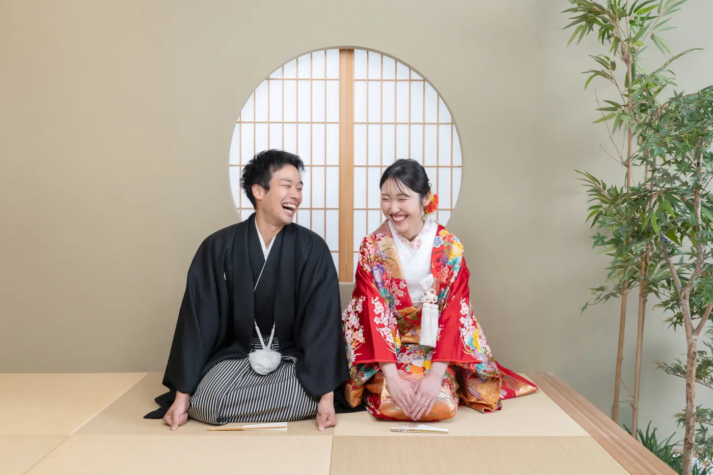丸窓のある和室で、顔を見合わせて笑う赤い色打掛の花嫁と黒紋付袴の花婿