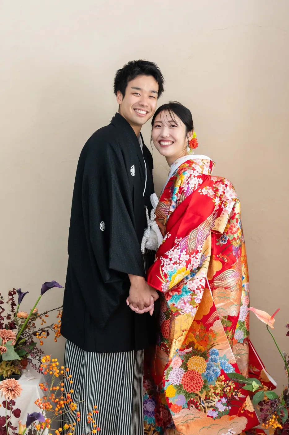 花に囲まれて手をつなぎ、笑顔で寄り添う赤い色打掛の花嫁と黒紋付袴の花婿