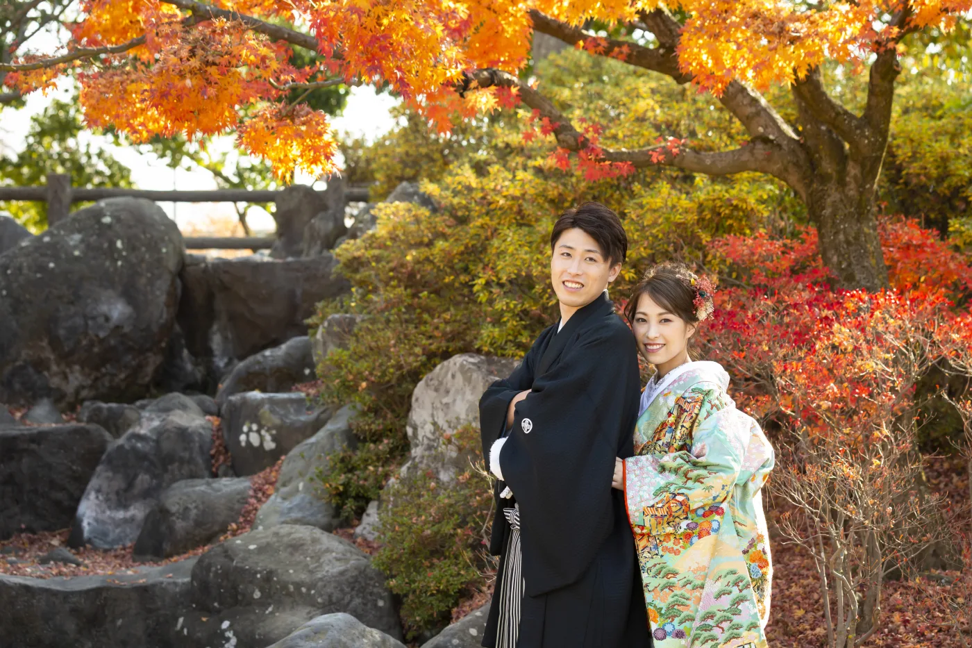 あがたの森の紅葉の下、岩を背に寄り添って立つ色打掛の花嫁と黒紋付袴の花婿