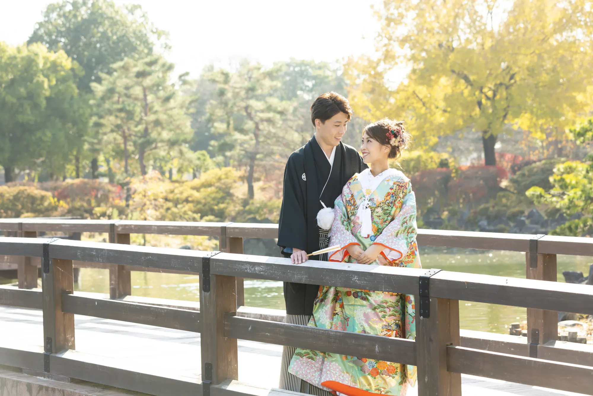池のほとりの木の橋の上で、見つめ合う水色の色打掛の花嫁と黒紋付袴の花婿