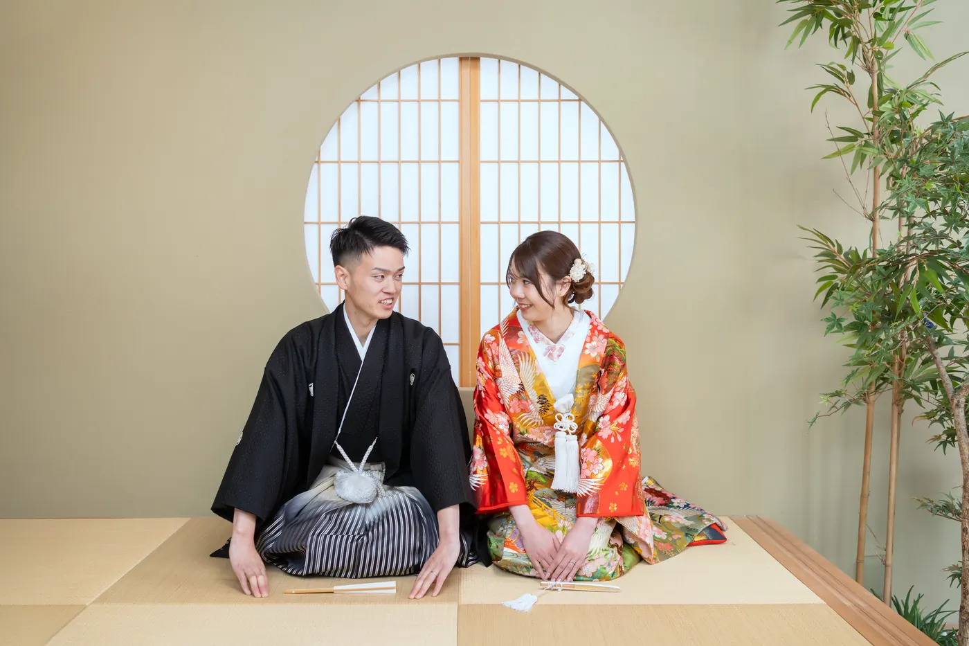 丸窓のある和室で並んで座る、赤い色打掛の花嫁と黒紋付袴の花婿