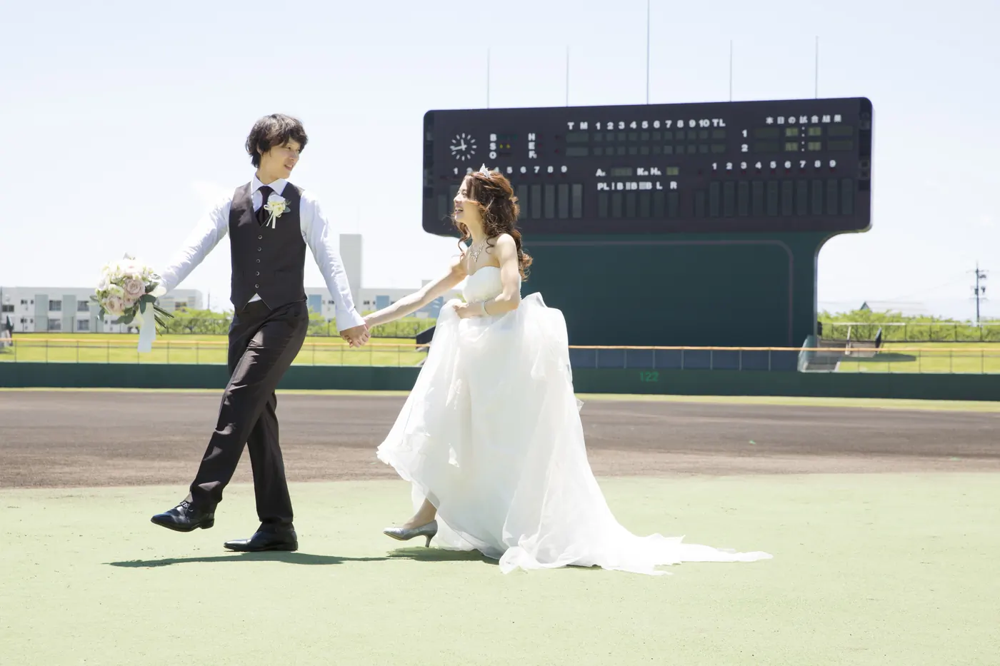 野球場のスコアボードの前で手をつなぐ、ドレスの花嫁とタキシードの花婿